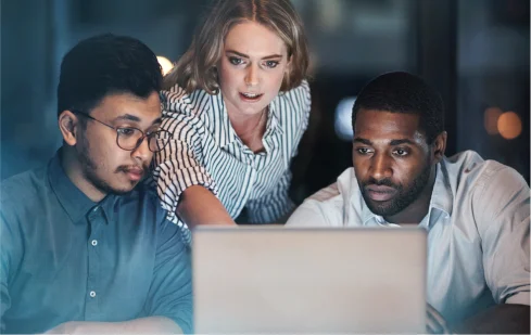 A team meeting at night with three professionals, two men and one woman, focusing intently on a laptop screen.